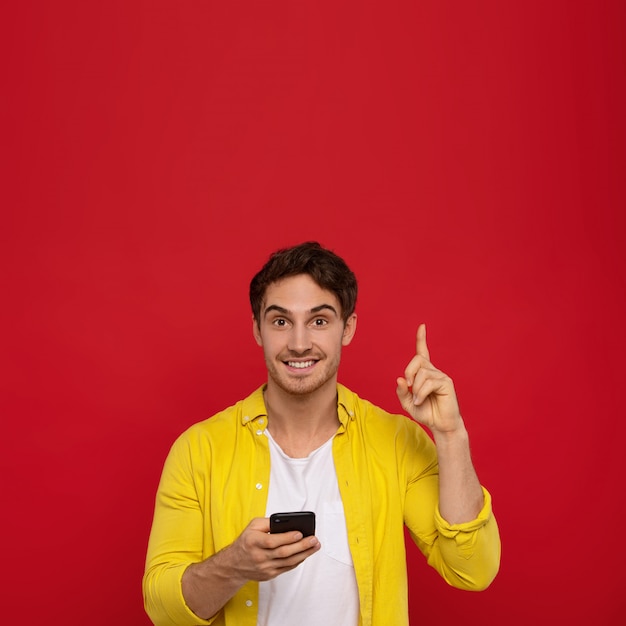 Hombre sonriente en camisa amarilla con teléfono inteligente y apuntando con el dedo hacia arriba, sonríe positivamente,