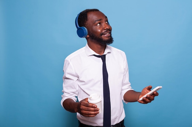 Hombre sonriente con camisa blanca mirando hacia arriba sosteniendo un teléfono inteligente transmitiendo música a auriculares inalámbricos mientras sostiene una taza de bebida caliente. Estudiante relajante con café para llevar navegando por las redes sociales.