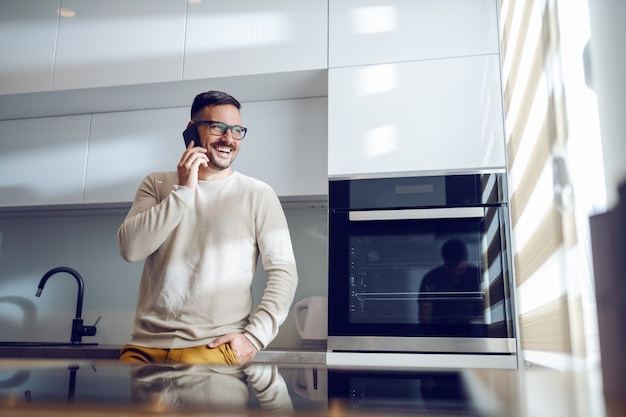 Hombre sonriente caucásico guapo en suéter apoyándose en la encimera y hablando por teléfono. Interior de cocina doméstica.