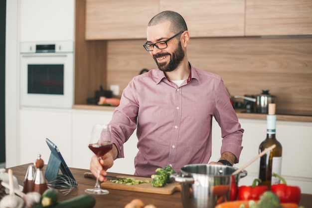 Hombre sonriente con copa de vino cocinando guiso de verduras con tableta. Está cortando brócoli, calabacín, pimiento rojo, cebolla y otras verduras.