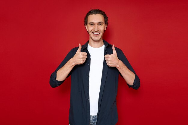 Hombre sonriente dando pulgares arriba signo de aprobación ropa casual de calidad aislado sobre fondo rojo.