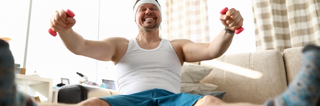 Hombre sonriente divertido sentado en fit ball trabajando con pequeñas pesas rosas en casa durante el período de cuarentena de coronavirus retrato