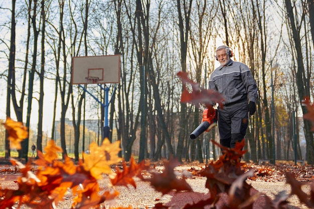 Hombre sonriente de edad trabajando con un soplador de hojas de mano mientras las hojas se rizan y brillan de manera agradable