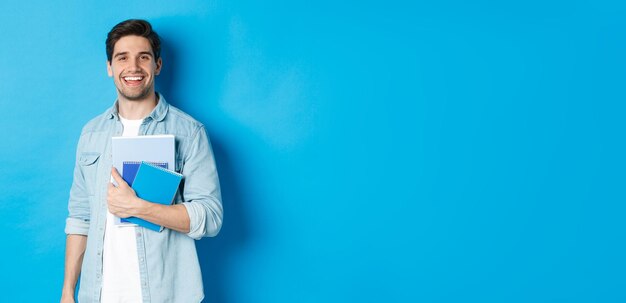 Foto hombre sonriente estudiando sosteniendo cuadernos y mirando feliz de pie sobre fondo azul.