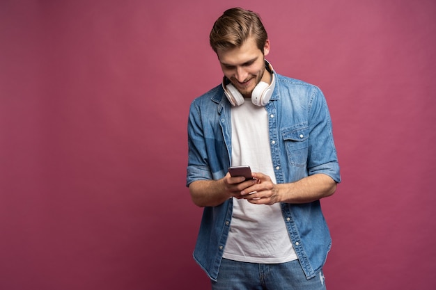 Hombre sonriente feliz con auriculares escuchando música en su teléfono inteligente aislado sobre fondo rosa. Estilo de vida de entretenimiento móvil