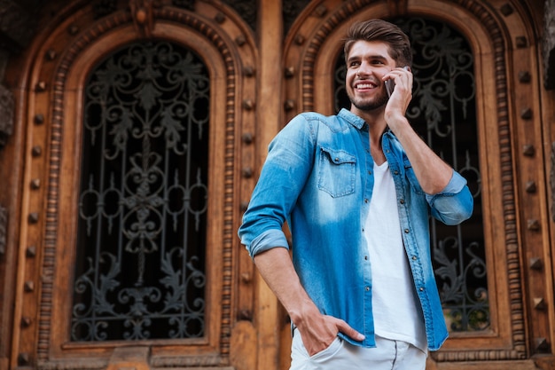 Hombre sonriente feliz hablando por el teléfono móvil mientras está de pie frente a la puerta de madera en la calle