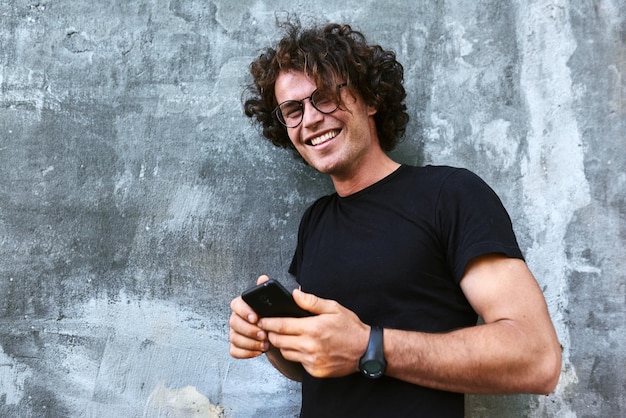 Hombre sonriente feliz de pie al aire libre enviando mensajes de texto en el teléfono móvil Hombre joven con cabello rizado usa gafas descansando afuera en la ciudad navegando en su teléfono celular sobre fondo gris concreto