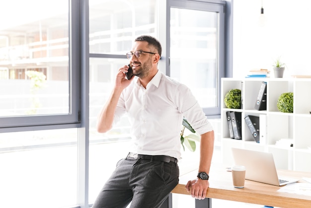 Hombre sonriente feliz en ropa formal trabajando y hablando en un teléfono inteligente negro, mientras está de pie cerca de una ventana grande en la oficina