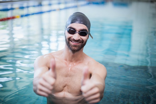 Hombre sonriente con gorra y gafas de natación con los pulgares arriba en la piscina