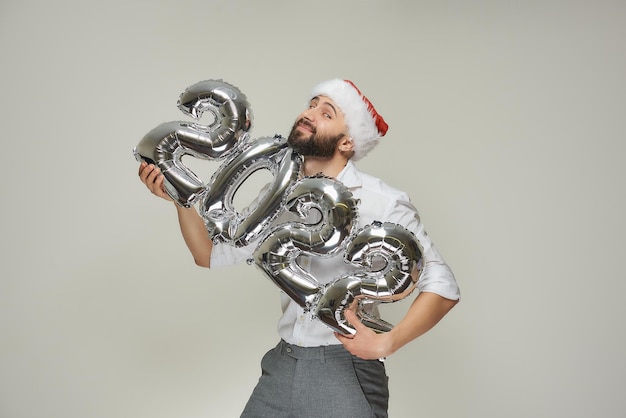 Un hombre sonriente con un gorro de Papá Noel de terciopelo rojo abraza suavemente globos plateados con la forma de 2022. Un chico feliz con barba en una fiesta de año nuevo.