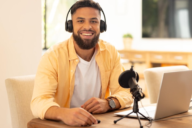 Hombre sonriente grabando podcast con micrófono y portátil en su oficina en casa