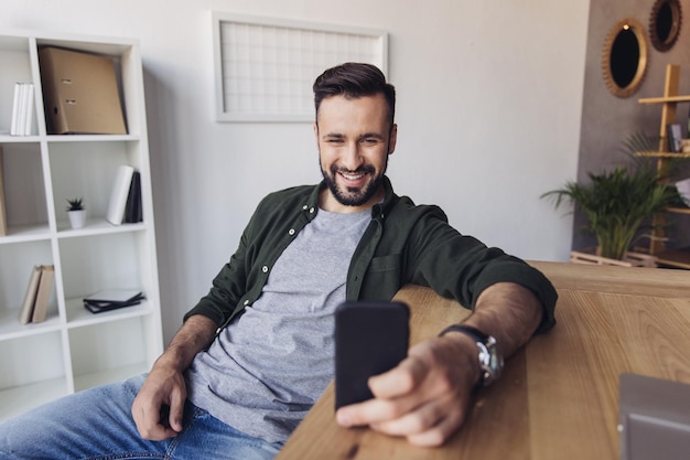 Foto hombre sonriente guapo usando un teléfono inteligente mientras está sentado en la mesa de la oficina