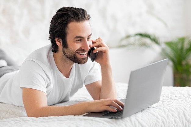 Hombre sonriente haciendo una llamada telefónica y usando una computadora portátil en la cama