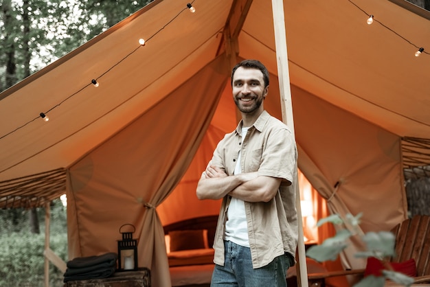 Hombre sonriente joven de pie dentro de la tienda glamping mirando la naturaleza del bosque. Cabina exterior de madera con bombillas. Acogedor, camping, glamping, vacaciones, concepto de estilo de vida de vacaciones.