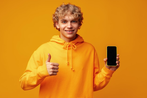 Hombre sonriente joven que sostiene el teléfono inteligente con la pantalla del dispositivo en blanco contra el fondo amarillo