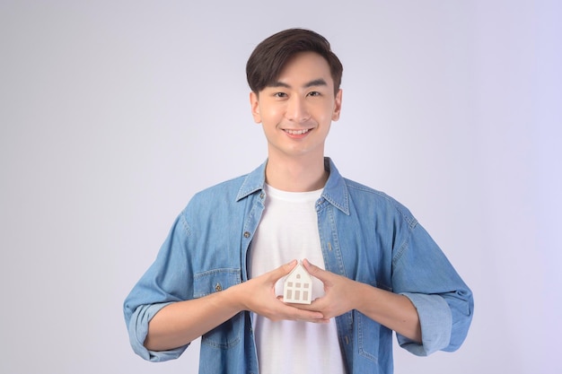 Hombre sonriente joven sosteniendo una casa modelo pequeña sobre fondo blanco studiox9