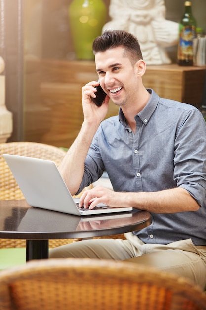 Foto hombre sonriente en llamada telefónica en el café con la computadora portátil