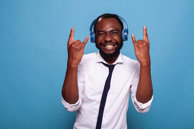 Hombre sonriente lleno de energía haciendo el signo de la mano de rock and roll mientras escucha música en auriculares inalámbricos. Hombre de negocios actuando rebelde haciendo señales con la mano mientras escucha música alta en auriculares.