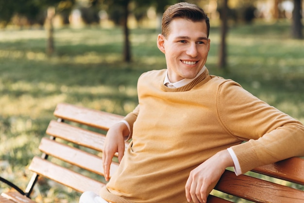 Hombre sonriente moderno y guapo con ropa amarilla y blanca sentado en un banco del parque al atardecer