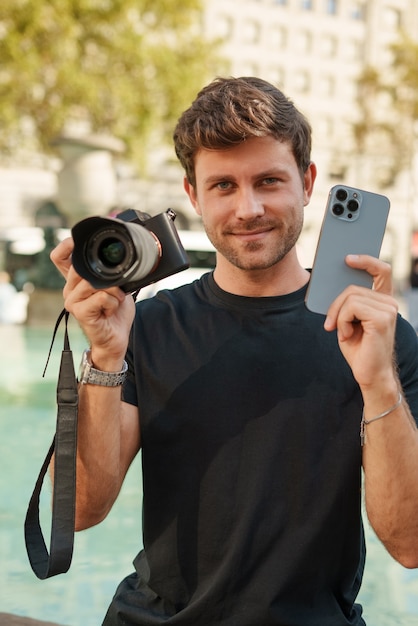 Hombre sonriente mostrando cámara de fotos y teléfono inteligente en la plaza de la ciudad