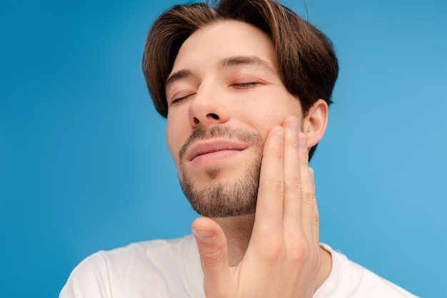 Hombre sonriente con los ojos cerrados tocando su cara con la mano posando ante la cámara en el estudio aislado sobre un fondo azul concepto de belleza de cuidado de la piel
