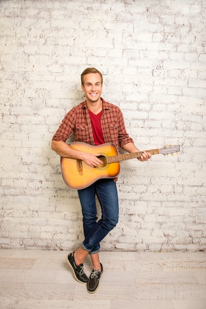 Hombre sonriente de pie cerca de la pared y tocando la guitarra