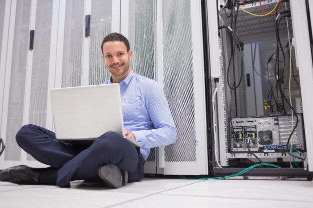 Hombre sonriente que se sienta en los servidores de comprobación del piso con la computadora portátil