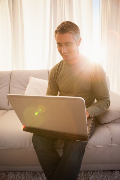 Hombre sonriente que se sienta en el sofá usando la computadora portátil