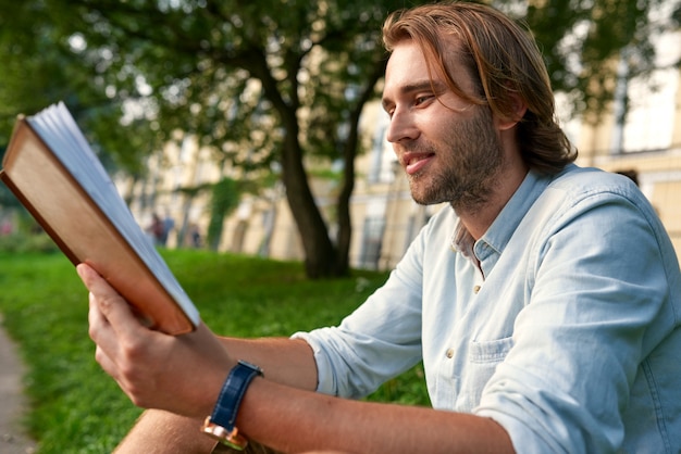 Foto hombre sonriente que sostiene un libro en la hierba en el campus.