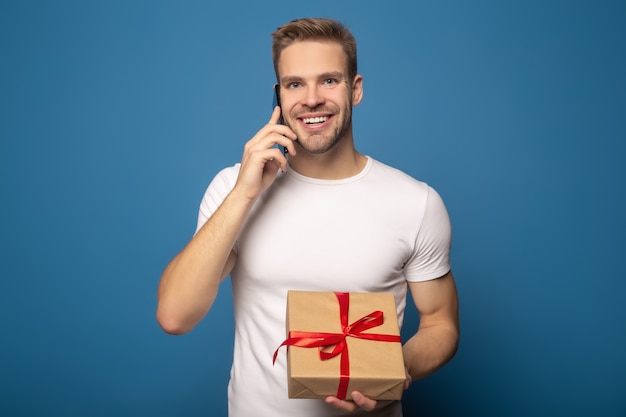 Hombre sonriente que sostiene el regalo y que habla en el teléfono inteligente aislado en azul