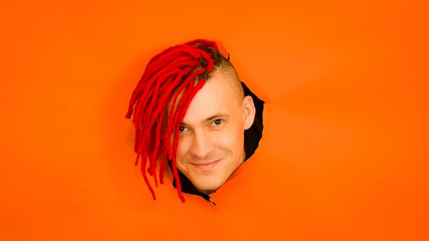 Foto hombre sonriente con rastas mirando a la cámara varón joven alegre con el pelo teñido mirando a la cámara en el agujero sobre fondo naranja en el estudio