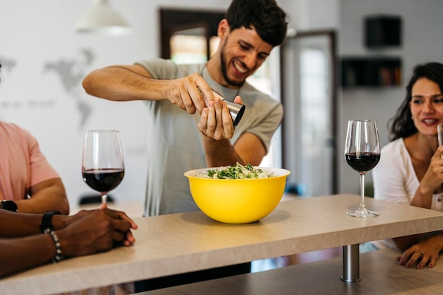 Hombre sonriente rociando sal en una ensalada entre amigos bebiendo vino tinto