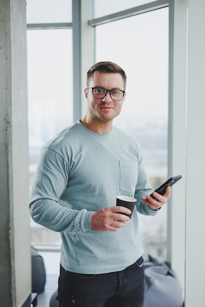 Hombre sonriente con ropa informal parado cerca de la ventana con café y mirando el teléfono inteligente en un espacio de trabajo moderno con una ventana grande durante el día