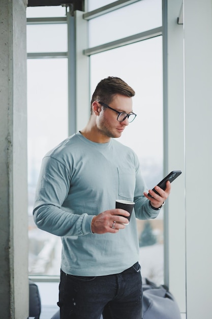 Hombre sonriente con ropa informal parado cerca de la ventana con café y mirando el teléfono inteligente en un espacio de trabajo moderno con una ventana grande durante el día