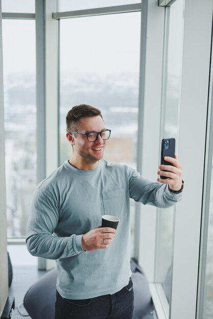 Hombre sonriente con ropa informal parado cerca de la ventana con café y mirando el teléfono inteligente en un espacio de trabajo moderno con una ventana grande durante el día