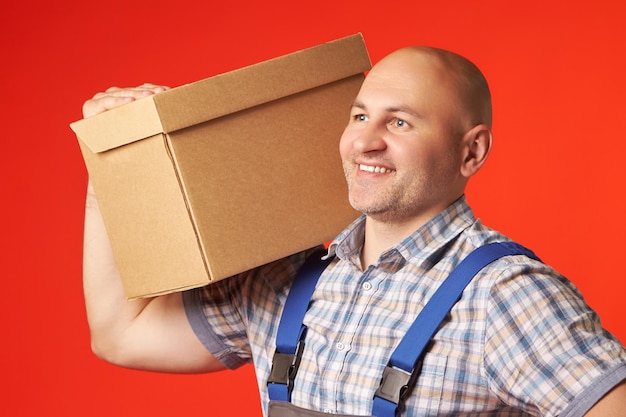 Hombre sonriente con ropa de trabajo sostiene una caja de cartón en el hombro y mira hacia otro lado