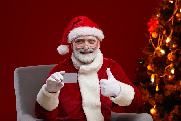 Hombre sonriente senior en traje de Santa sosteniendo la tarjeta de visita y mostrando el pulgar hacia arriba sentado en un sillón