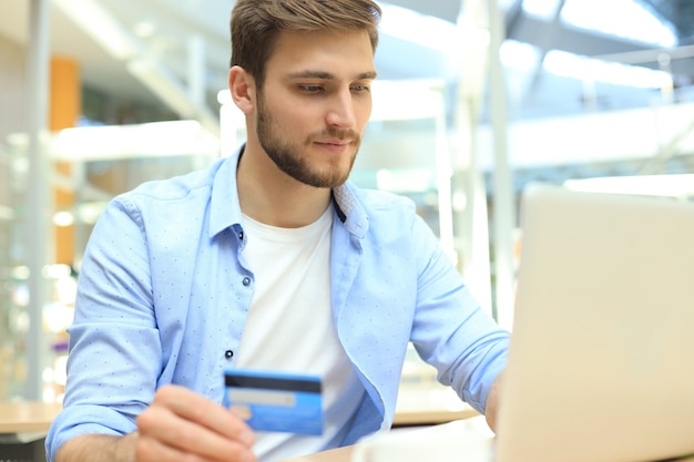 Hombre sonriente sentado en la oficina y paga con tarjeta de crédito con su computadora portátil.