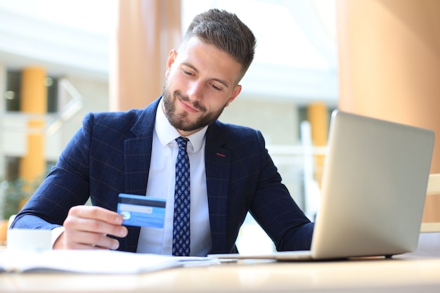 Foto hombre sonriente sentado en la oficina y paga con tarjeta de crédito con su computadora portátil.