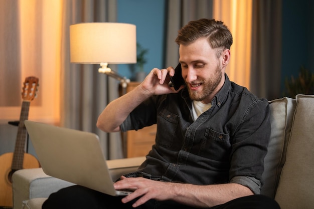 El hombre sonriente se sienta en la sala de estar en el sofá con una computadora portátil en las piernas trabajando remotamente ejecutando informes