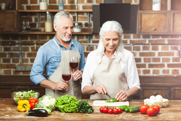 Hombre sonriente sosteniendo copas con vino tinto mientras la mujer prepara ensalada en la cocina