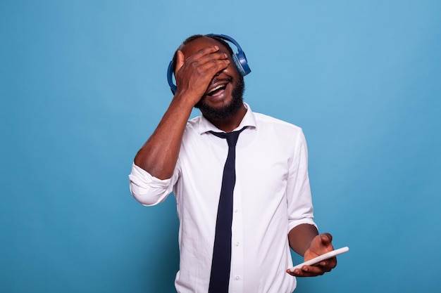 Hombre sonriente sosteniendo un teléfono inteligente que cubre los ojos con la palma de la mano para seleccionar una canción al azar de la lista de reproducción para escuchar en auriculares inalámbricos. Empresario escuchando un podcast divertido en auriculares con palmas en la cara.