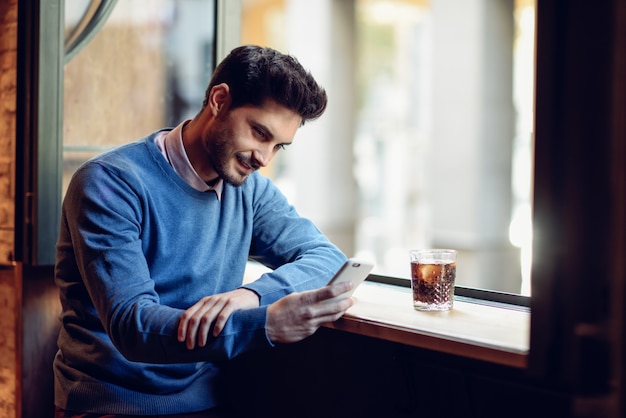 Foto hombre sonriente con suéter azul mirando su teléfono inteligente en un pub moderno.