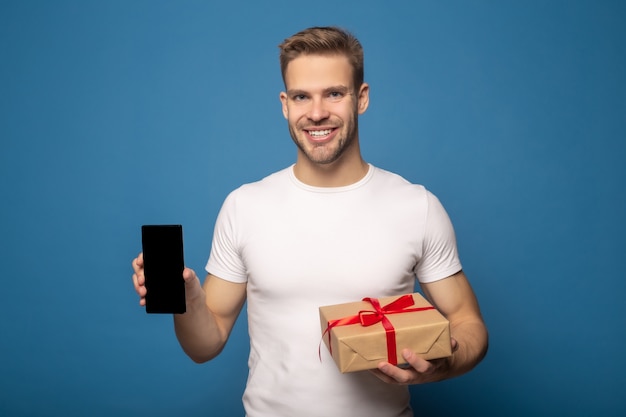 Hombre sonriente con teléfono inteligente y caja de regalo aislado en azul