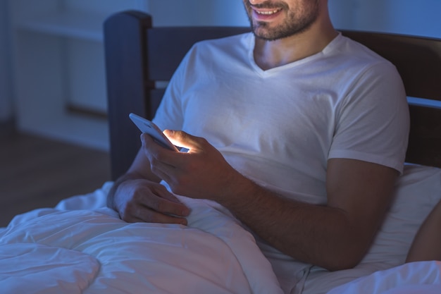 El hombre de la sonrisa se sienta con un teléfono en la cama. Noche