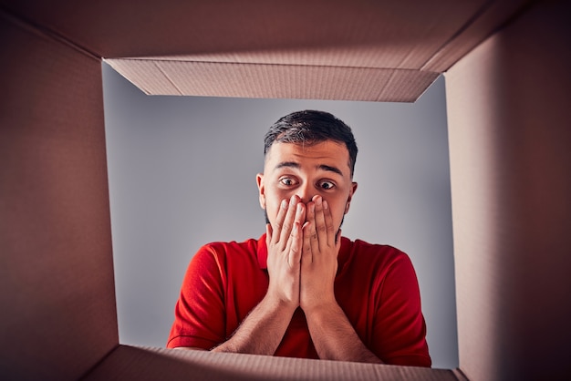 Foto el hombre sorprendido abriendo la caja de cartón y mirando dentro. el paquete, entrega, sorpresa, regalo, concepto de estilo de vida. conceptos de emociones humanas y expresiones faciales