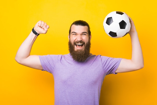 Hombre sorprendido con barba gritando y celebrando la victoria, fan apoyando a su equipo favorito y sosteniendo un balón de fútbol