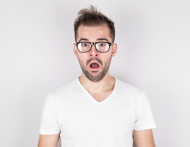 Hombre sorprendido con cabello despeinado con gafas en camiseta blanca en pared gris