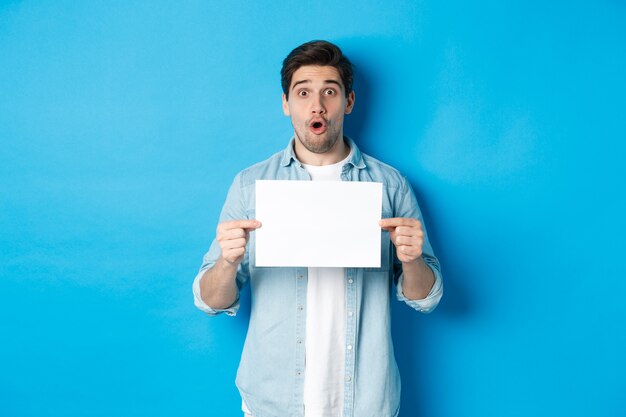 Foto hombre sorprendido jadeando y mirando impresionado a la cámara, mostrando un trozo de papel en blanco para su cartel, de pie sobre fondo azul