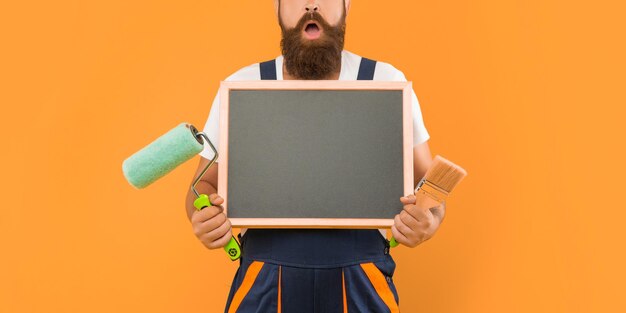 Hombre sorprendido recortado en ropa de trabajo sostiene pincel de rodillo de pintura y pizarra con espacio de copia